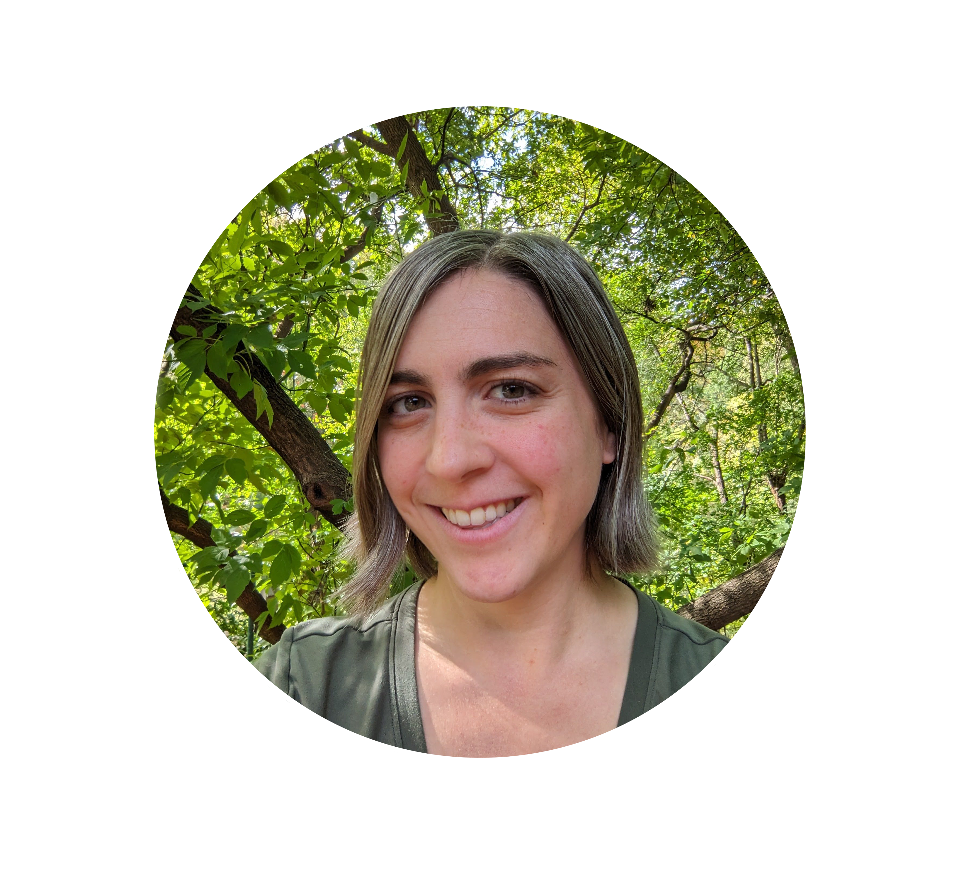 A color photo of Kris Tomes. Kris is looking at the viewer and her head and shoulders are in view. She has chin length hair, a light skin tone, and she is smiling, showing her top row of teeth. Branches and leaves are present in the background.